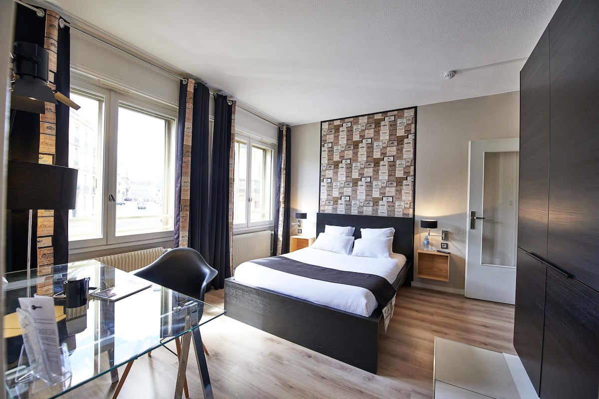 A modern hotel room with a glass desk, a bed with white linens, a dark headboard, and a wall decorated with framed pictures.