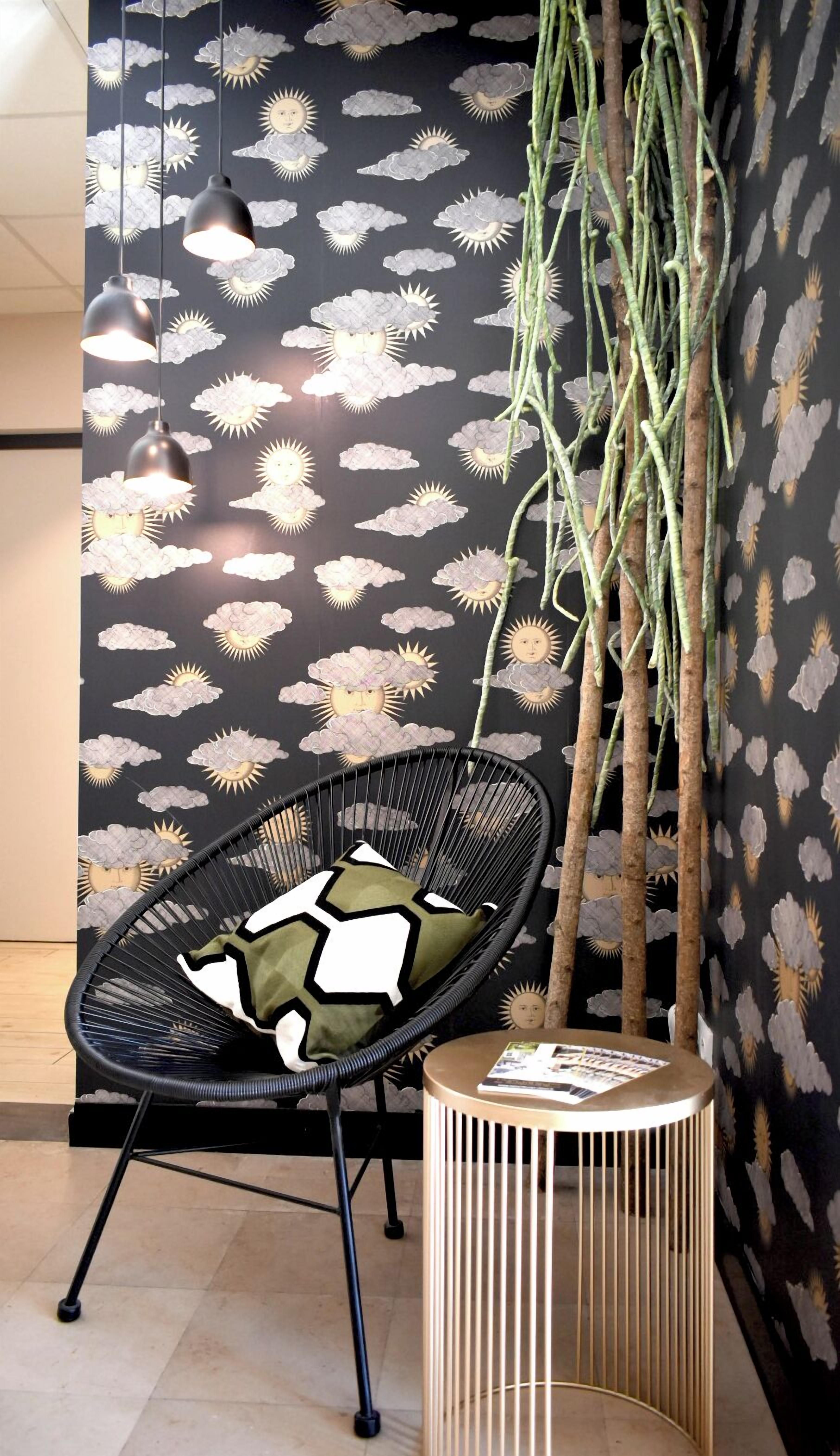 A room with a black wall featuring a weather pattern wallpaper, a black wire chair with a geometric pillow, a hanging plant, and a small wooden side table with a magazine.