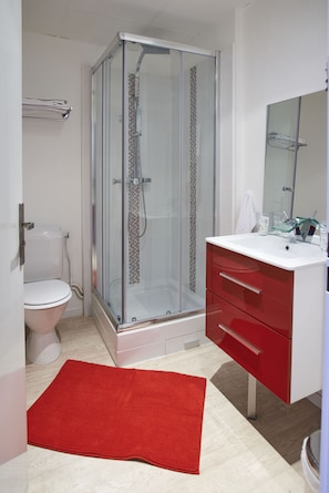 Un baño con ducha de vidrio, inodoro blanco, tocador rojo y alfombra roja.