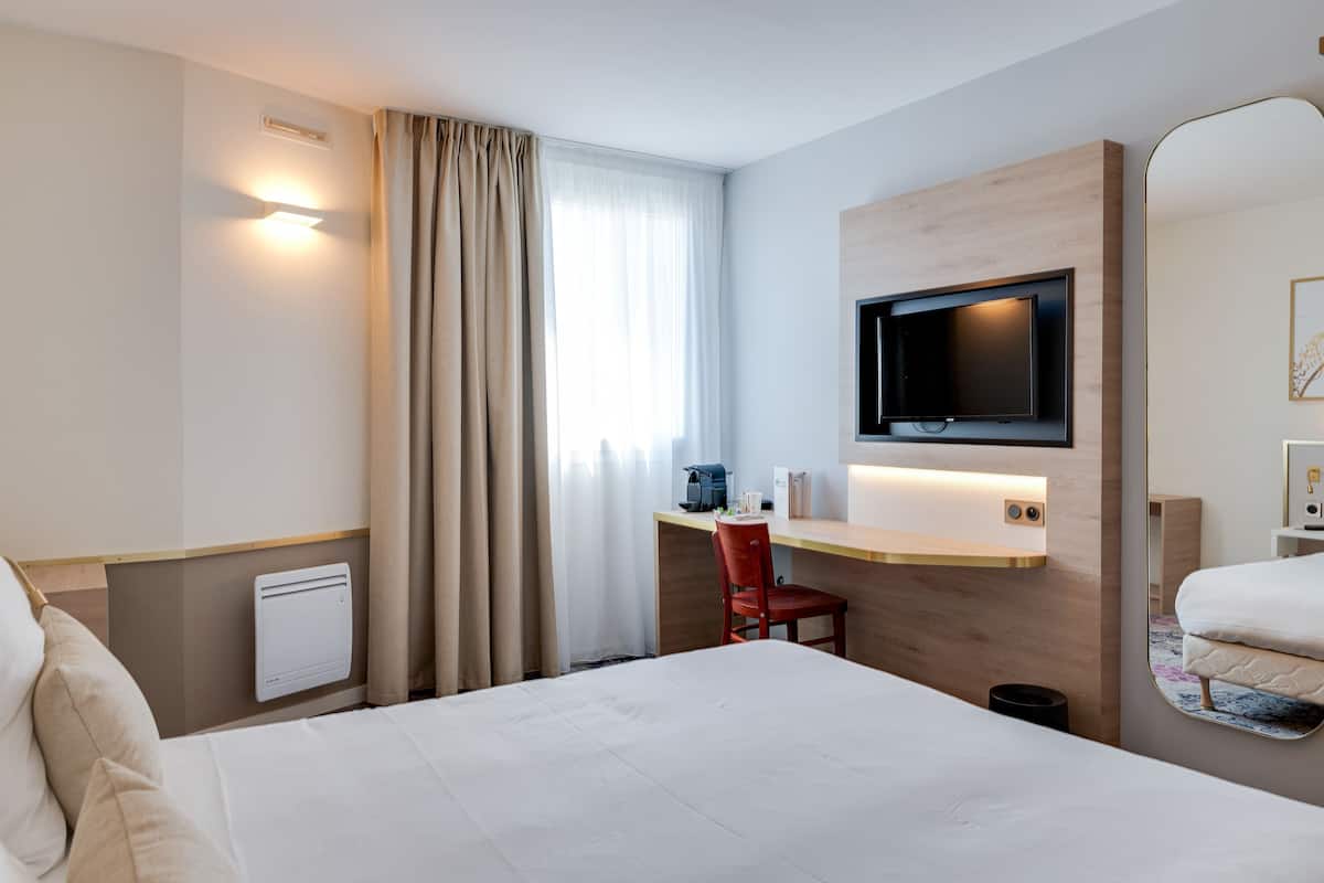 Une chambre d’hôtel comprenant un lit, un bureau avec une chaise, un téléviseur fixé au mur, un miroir et une fenêtre avec des rideaux.