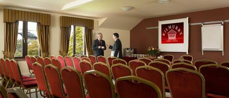 Un salón de conferencias con filas de sillas rojas, un atril y una pantalla proyectora mostrando 'Kilmurry Lodge Hotel'.