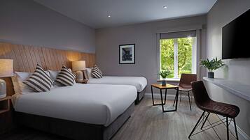 A modern hotel room with two beds, a small table, a chair, a TV, and a window with a view of greenery.