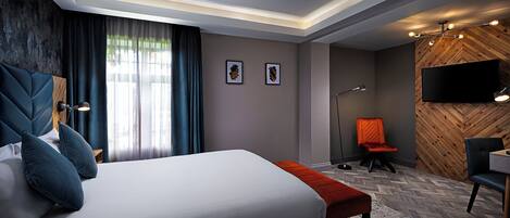 A modern hotel room with a large bed, a desk, a chair, a TV, and a lamp.