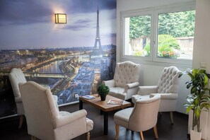 Una habitación con un gran mural de la Torre Eiffel, una ventana con vista a follaje, y un rincón de descanso con una mesa y sillas.