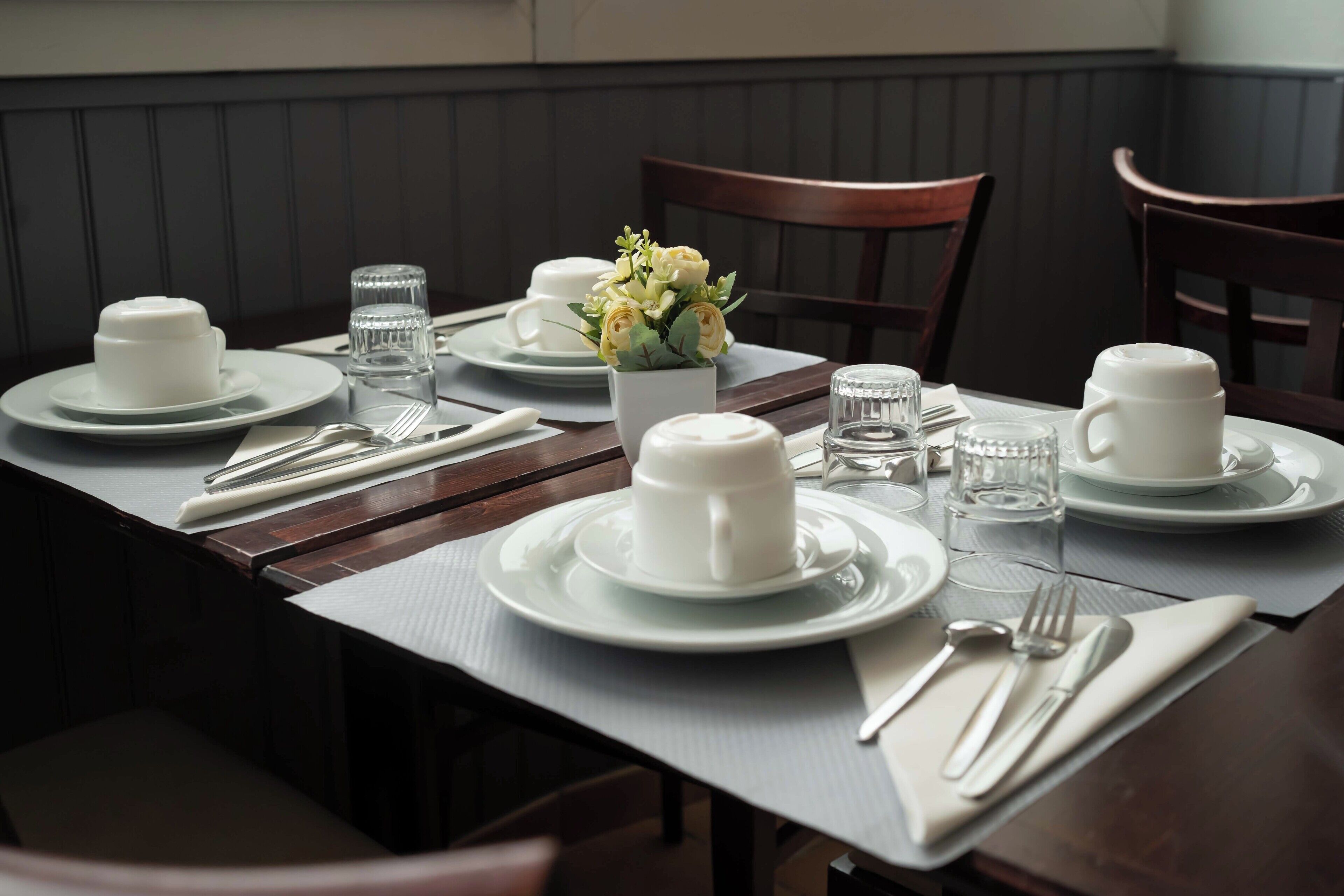 A neatly set dining table with white plates, cups, and cutlery, a vase of flowers, and a water glass.