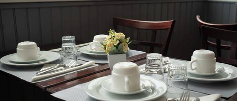 A neatly set dining table with white plates, cups, and cutlery, a vase of flowers, and a water glass.