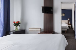 Une chambre d’hôtel avec un lit, un téléviseur fixé au mur, une table de chevet, un vase avec des fleurs et une porte donnant sur une autre pièce.