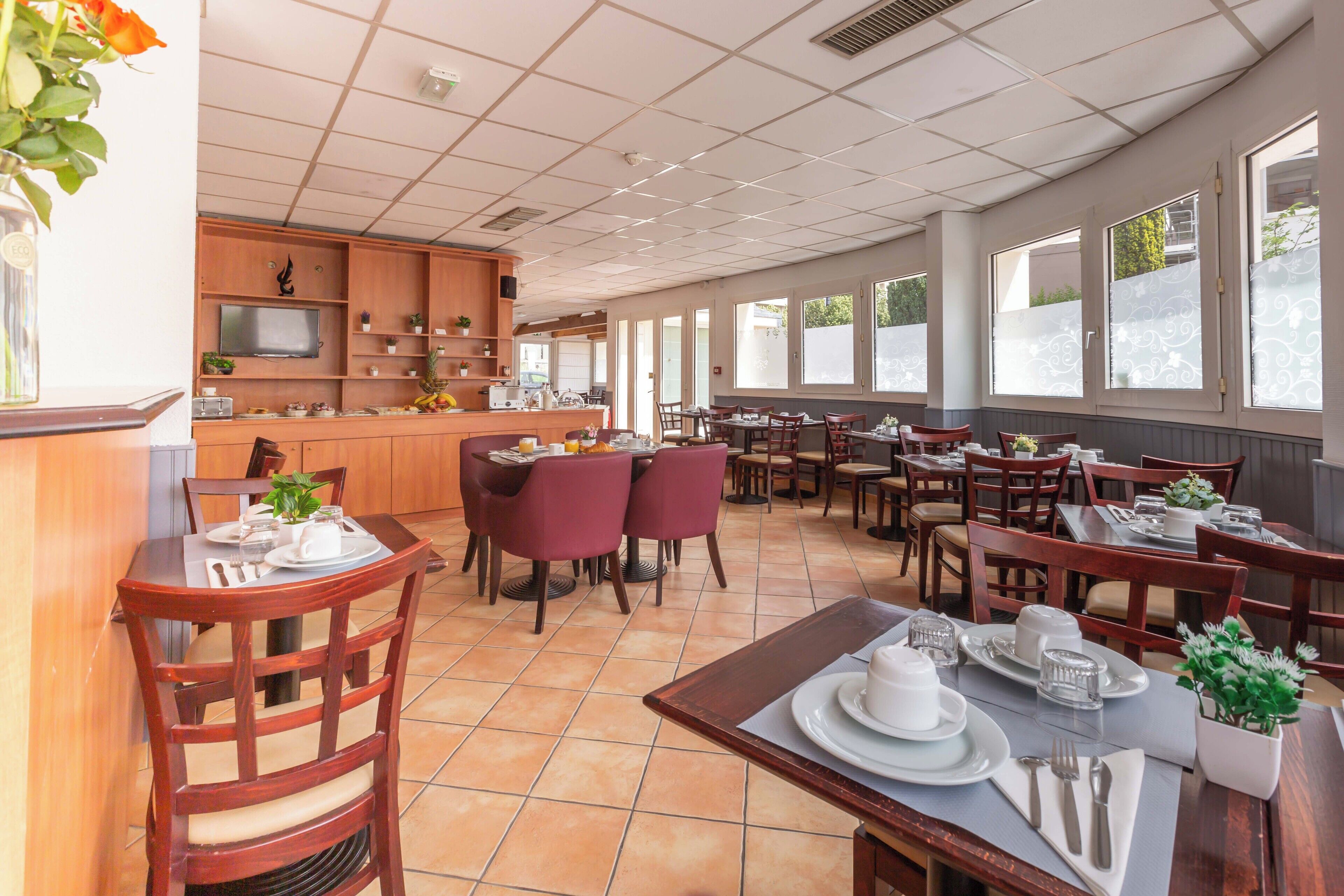 Un grand espace à manger avec des tables et des chaises en bois, un coin cuisine équipé d’un téléviseur, et de grandes fenêtres laissant entrer la lumière naturelle.