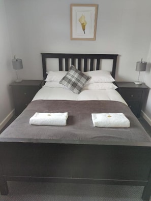 A neatly made bed with a brown bedspread, two white towels, and a plaid pillow. There are two bedside tables with lamps, and a framed picture on the wall above the bed.