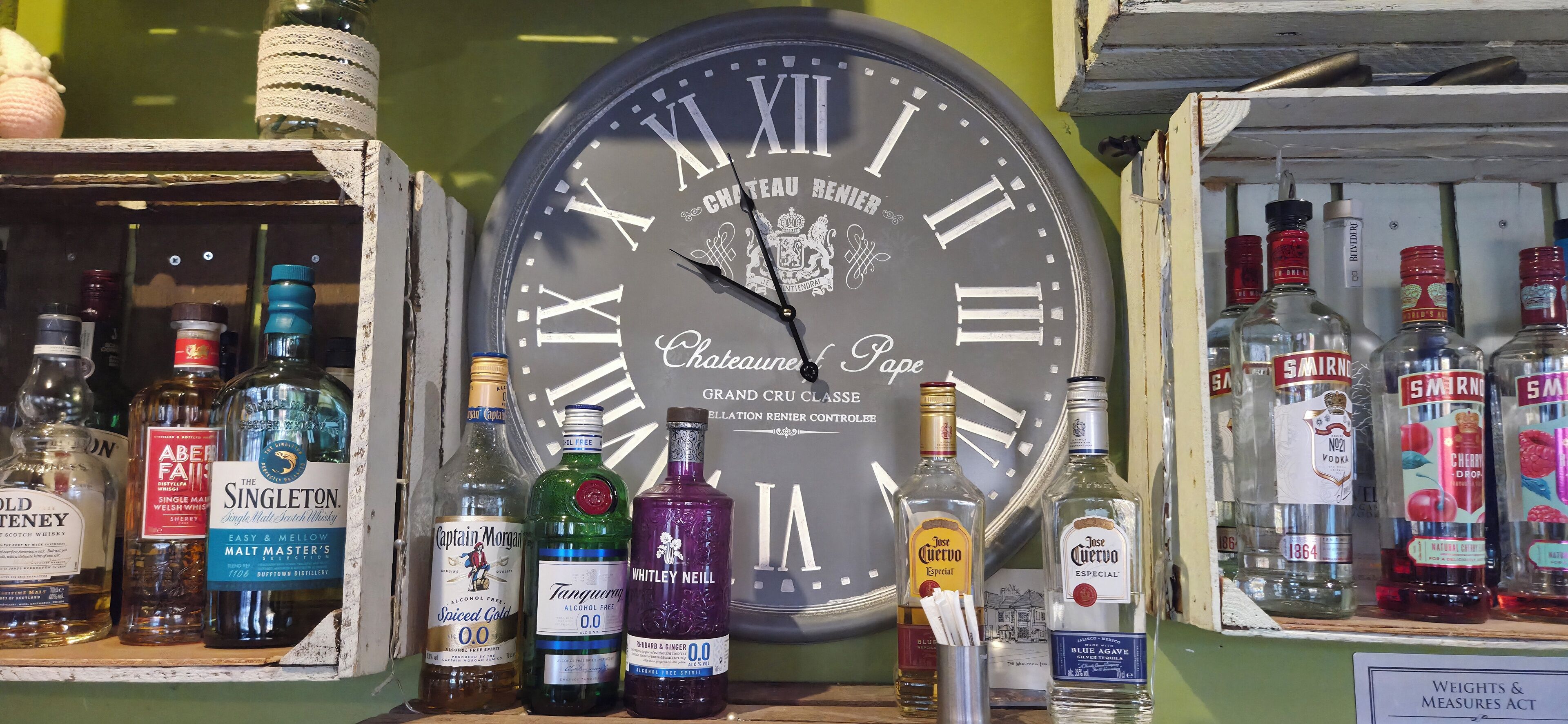Un reloj de pared con números romanos y la inscripción 'Chateau Renier', rodeado de varias botellas de licor.