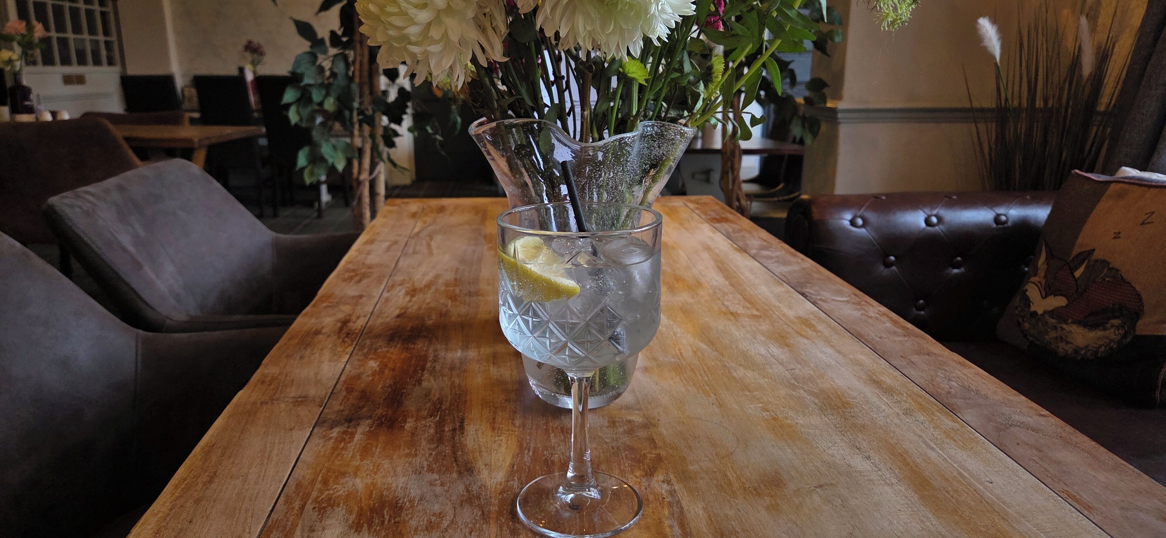 A clear glass with a lime wedge on a wooden table in a room with leather furniture and a vase of flowers.