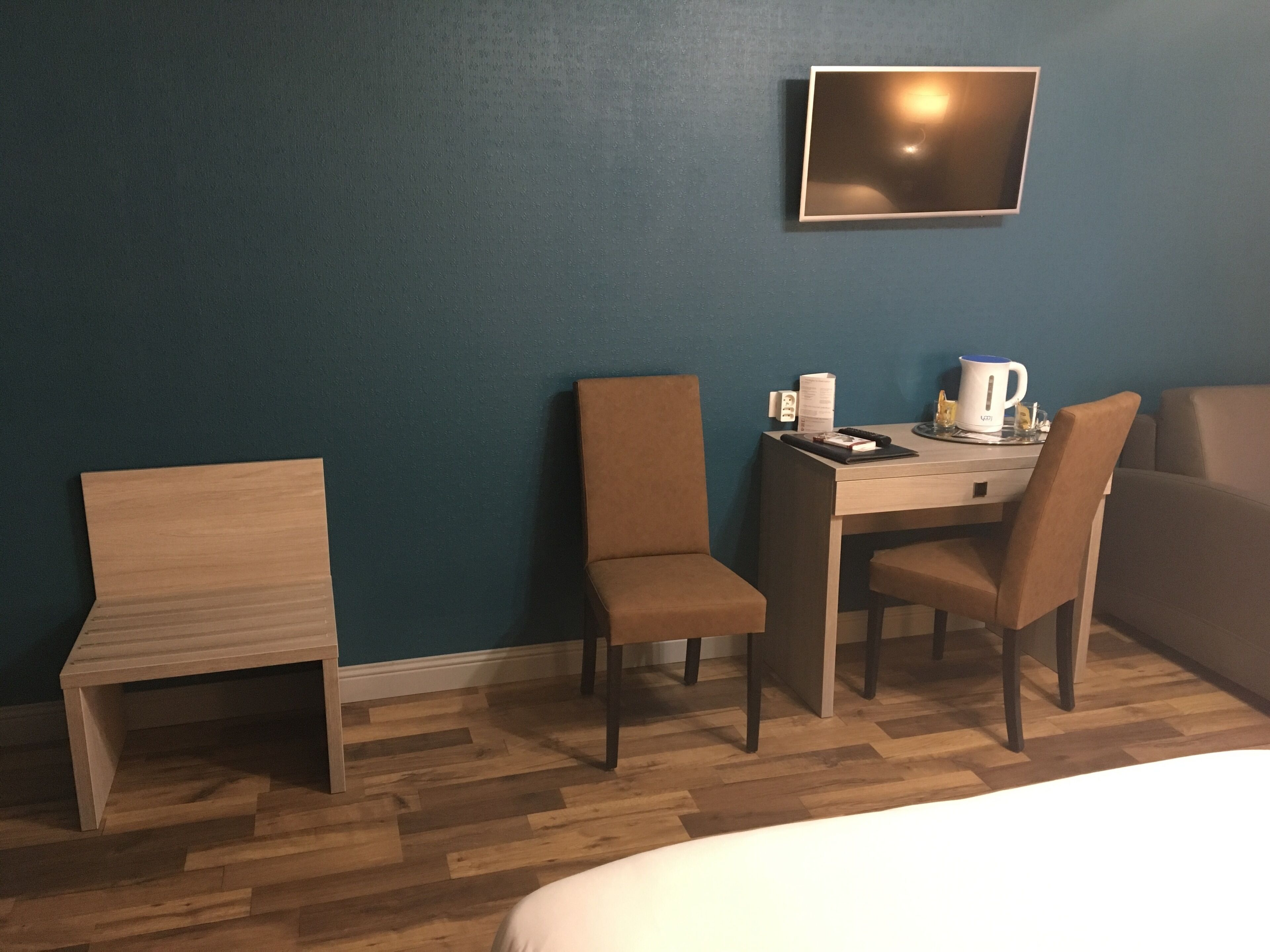 Quarto de hotel com uma cama, uma escrivaninha com duas cadeiras, uma cafeteira e uma televisão de tela plana na parede.