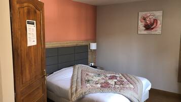 A hotel room with a wooden door, a bed with a floral quilt, a nightstand with a lamp, and a painting on the wall.