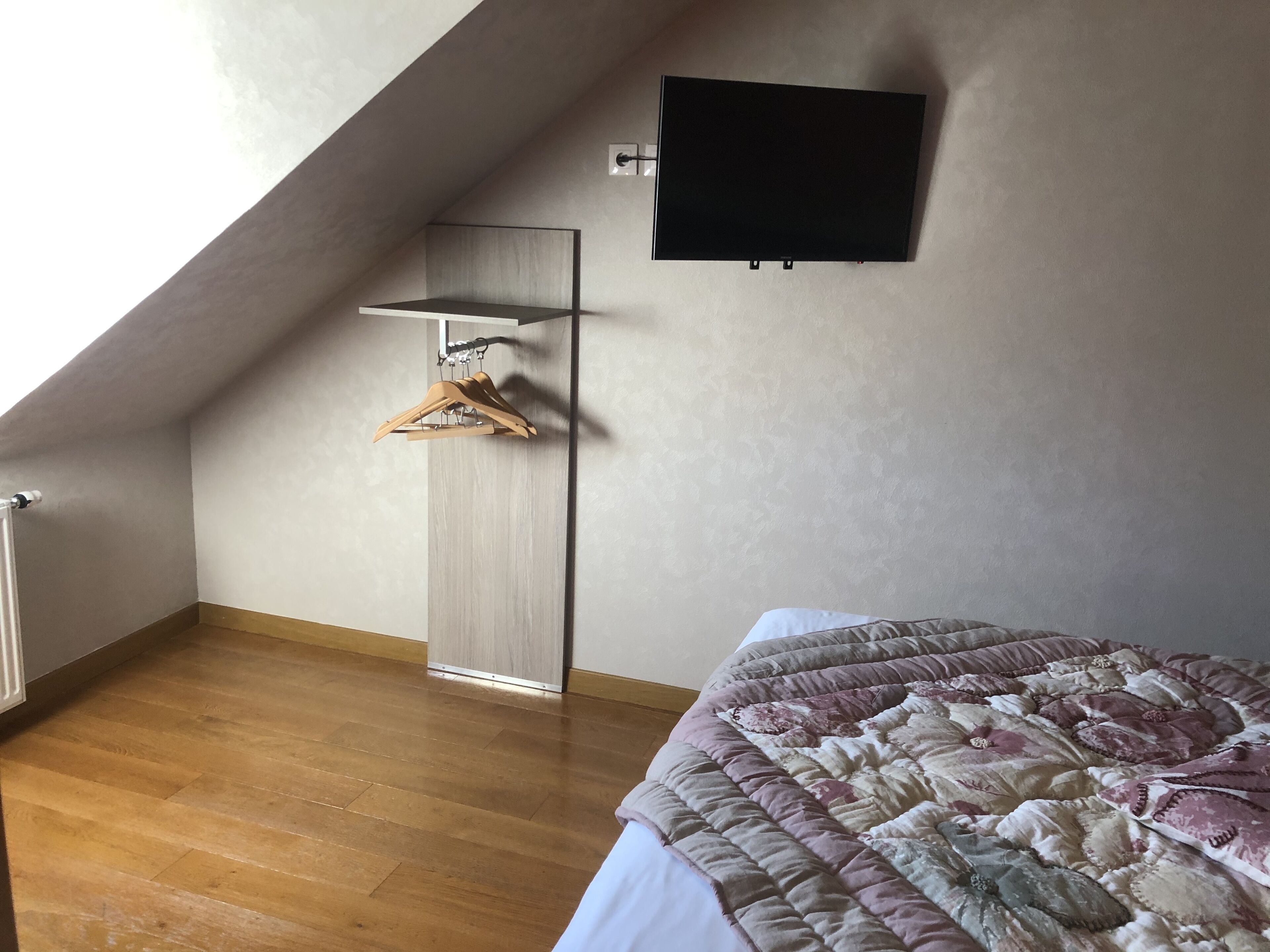 A bedroom with a sloped ceiling, a mounted TV, a wooden hanger rack, and a bed with a floral-patterned duvet.