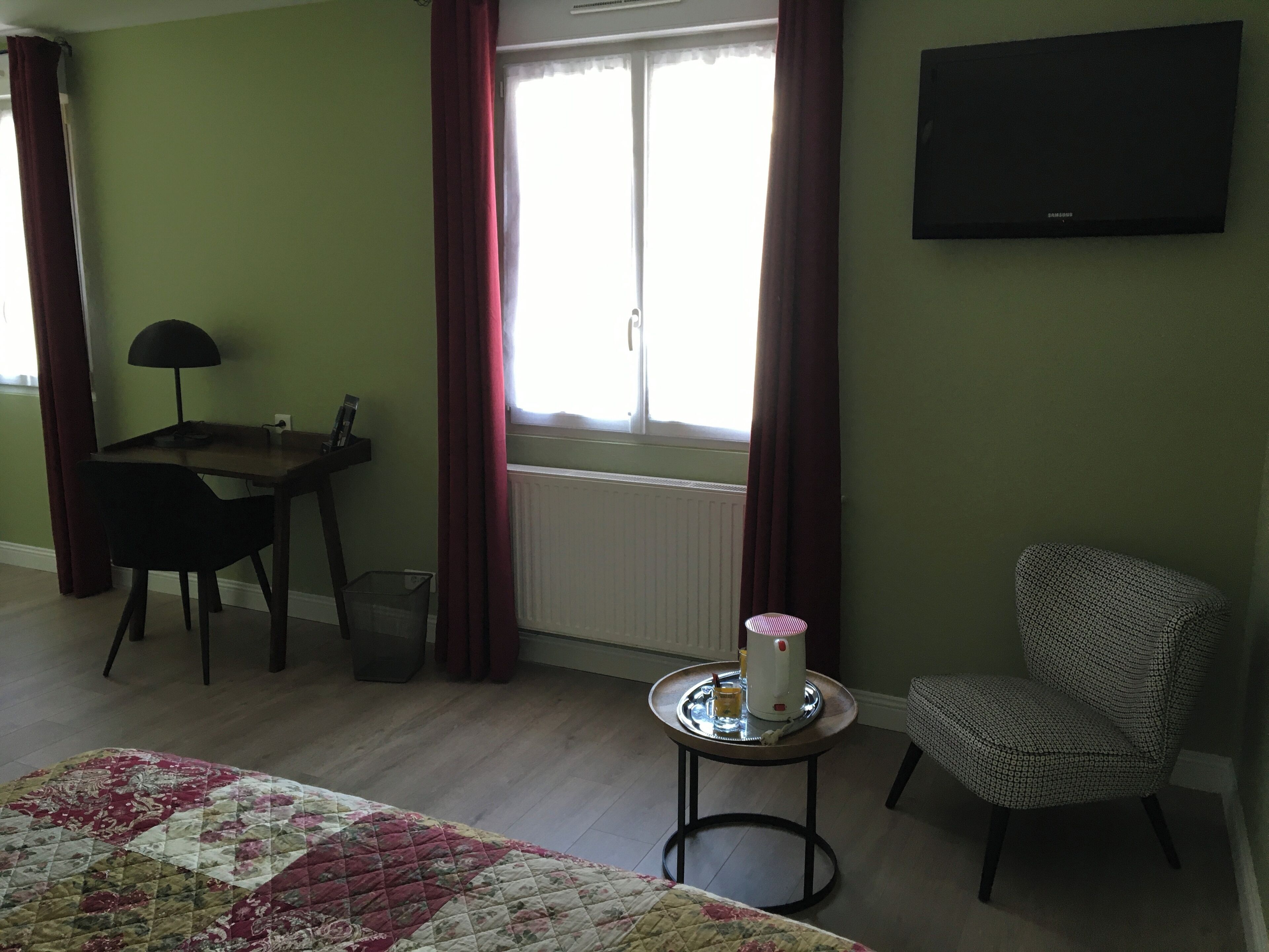 A room with a bed, a desk, a chair, a table with a kettle, and a TV on the wall.