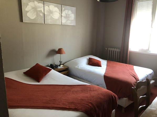 A hotel room with two beds, each with a red blanket and pillow, a nightstand with a lamp, and a wall art piece above the beds.