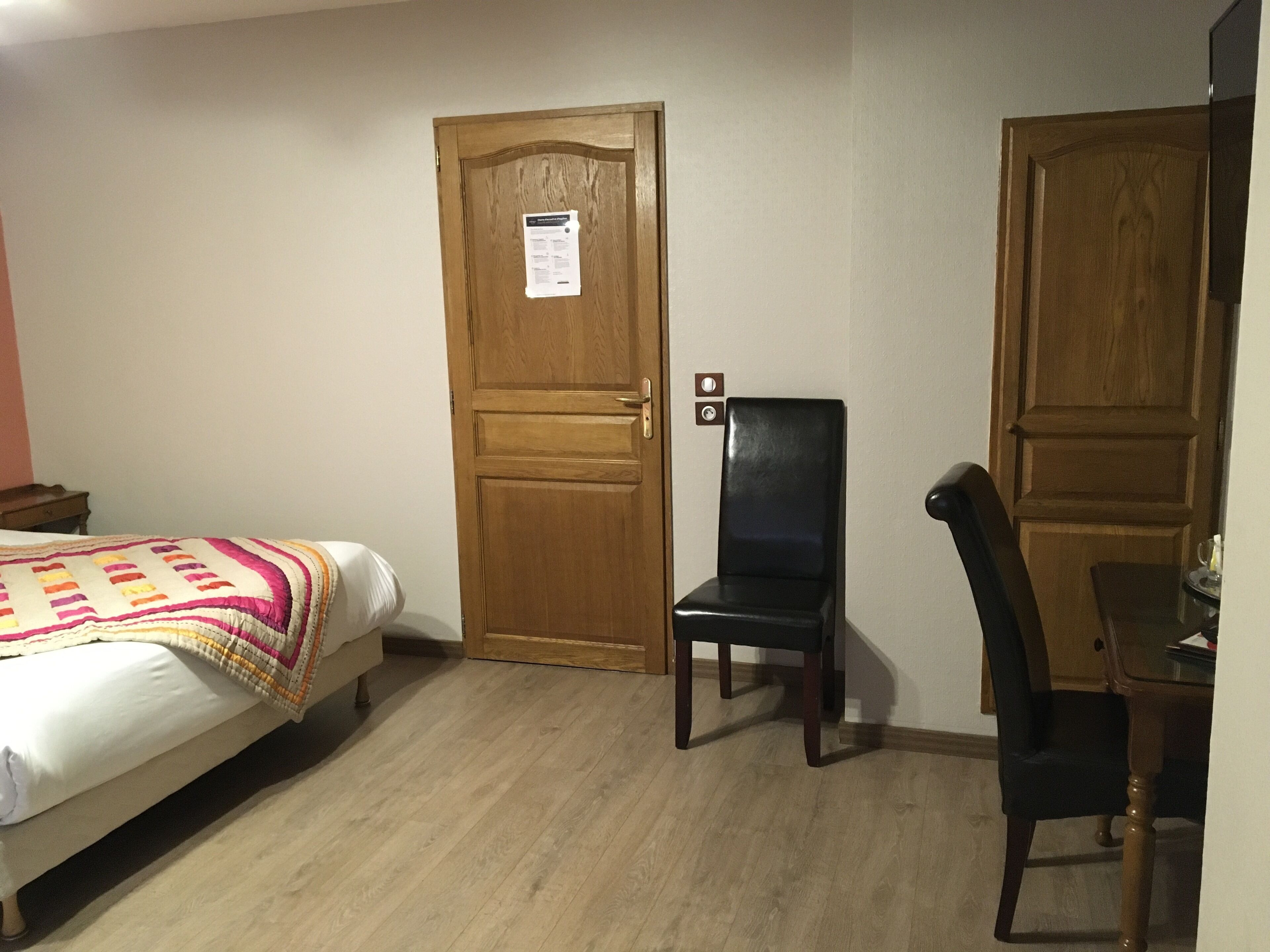 Ein Zimmer mit einem Bett, einer Holztür, einem schwarzen Stuhl und einem kleinen Tisch mit einem Glas.