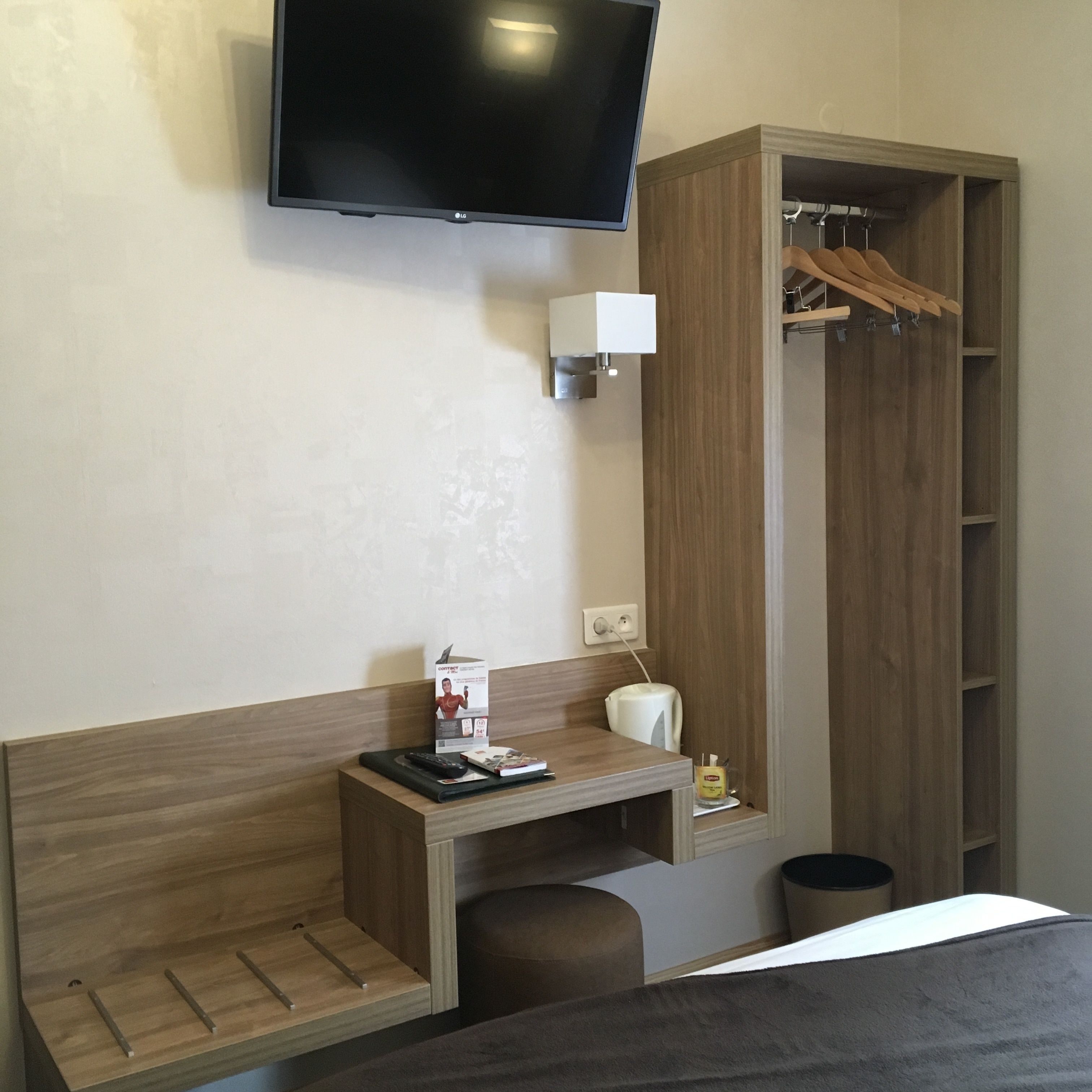 Habitación de hotel con televisor de pantalla plana montado en la pared, un escritorio de madera con un control remoto, un pequeño cesto de basura y un armario de madera con perchero.