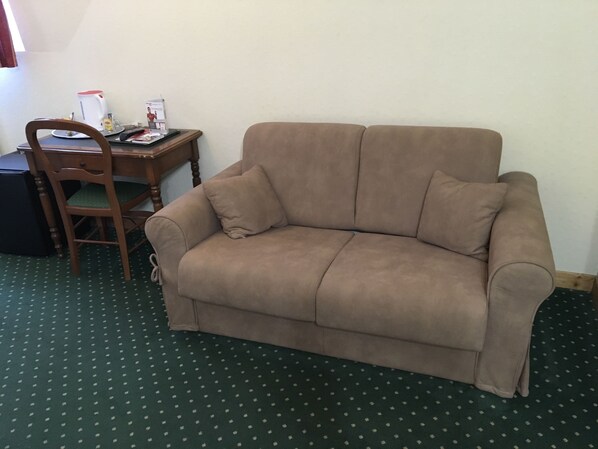 A beige sofa, a wooden chair, a small table with a kettle, and a patterned carpet.