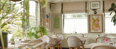 A dining area with wicker chairs, white tablecloths, and a window with curtains.