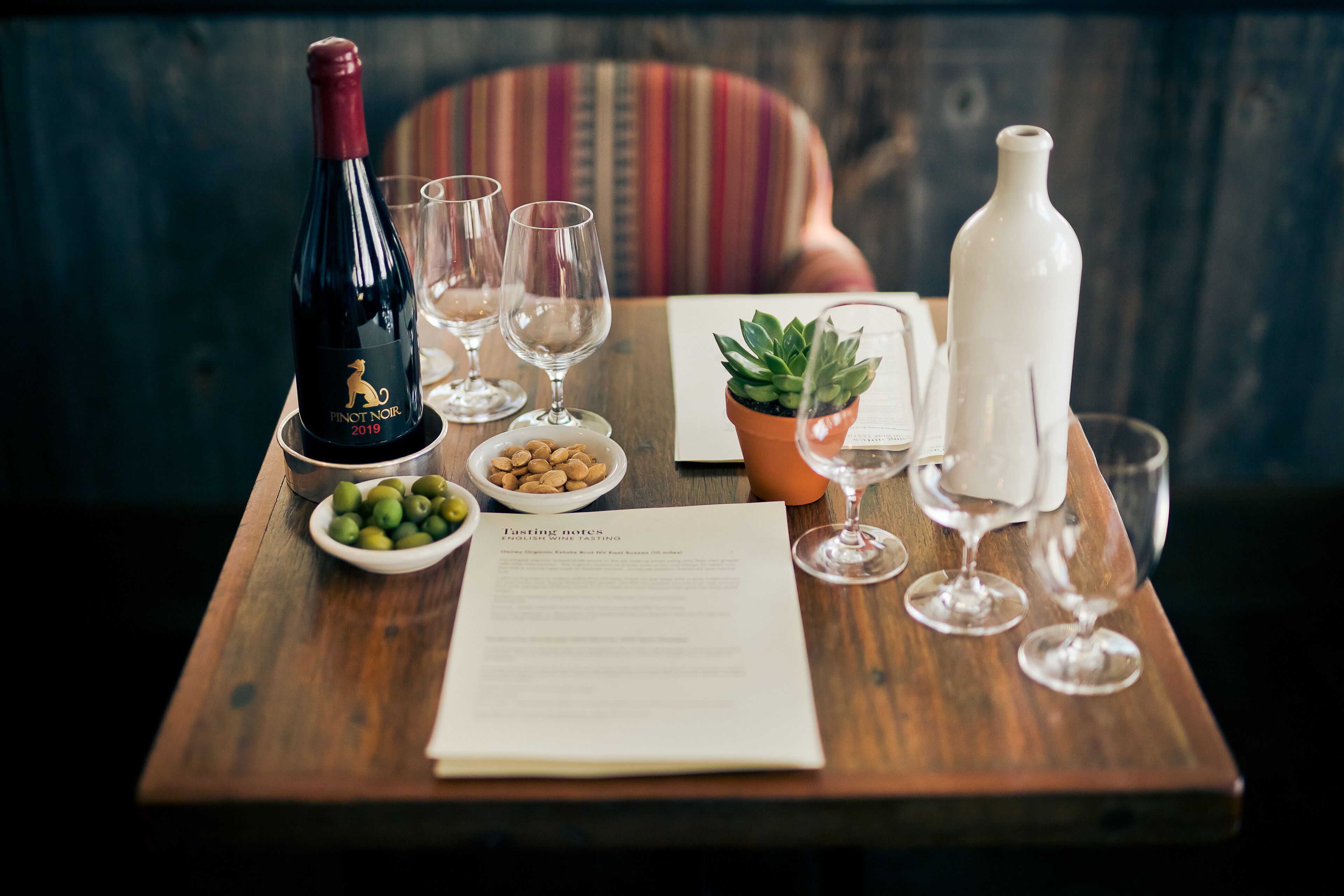 Ein Holztisch mit einer Flasche Pinot Noir, zwei Gläsern, einer Oliven-Schale, einem kleinen Topfpflänzchen, einer Speisekarte und einer weißen Flasche.