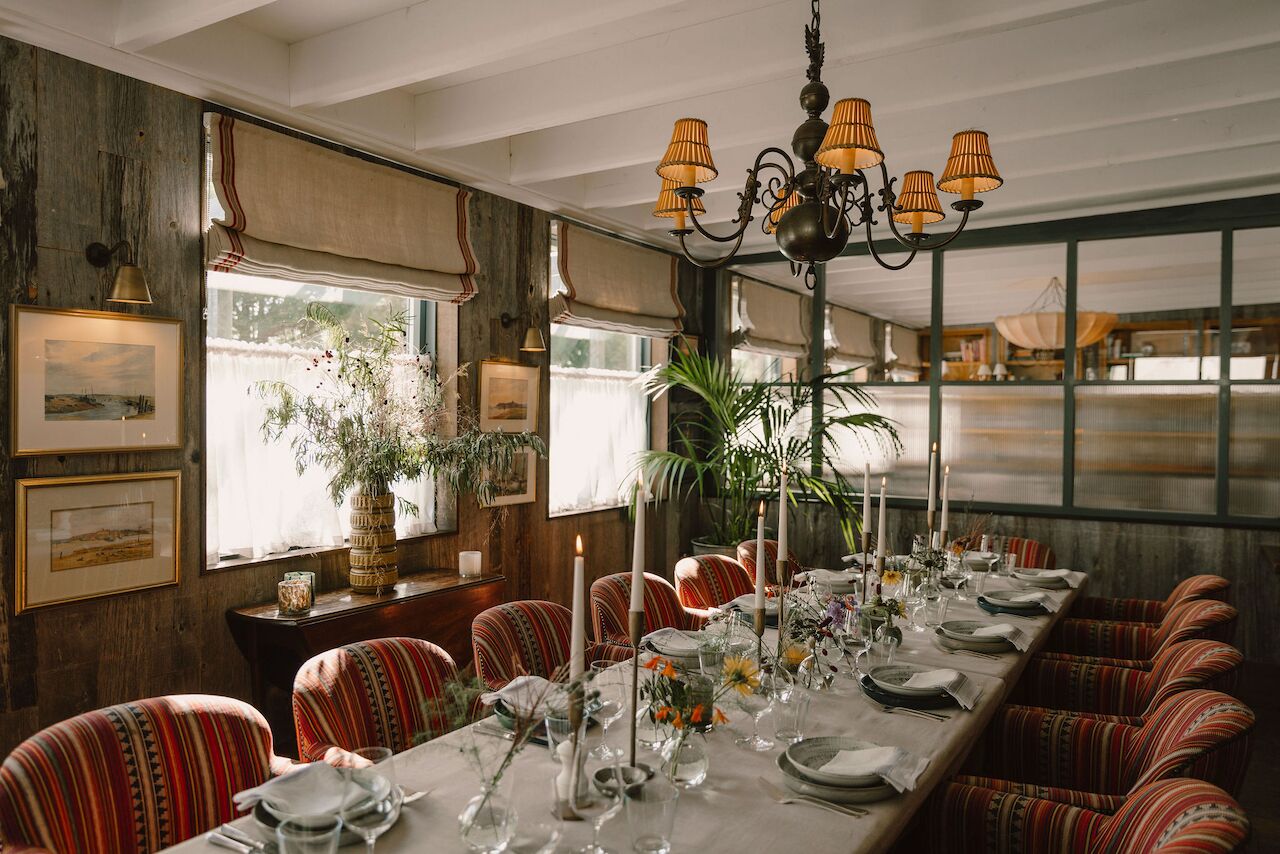 A long dining table set with candles, glassware, and floral centerpieces in a room with striped chairs, a chandelier, and large windows.
