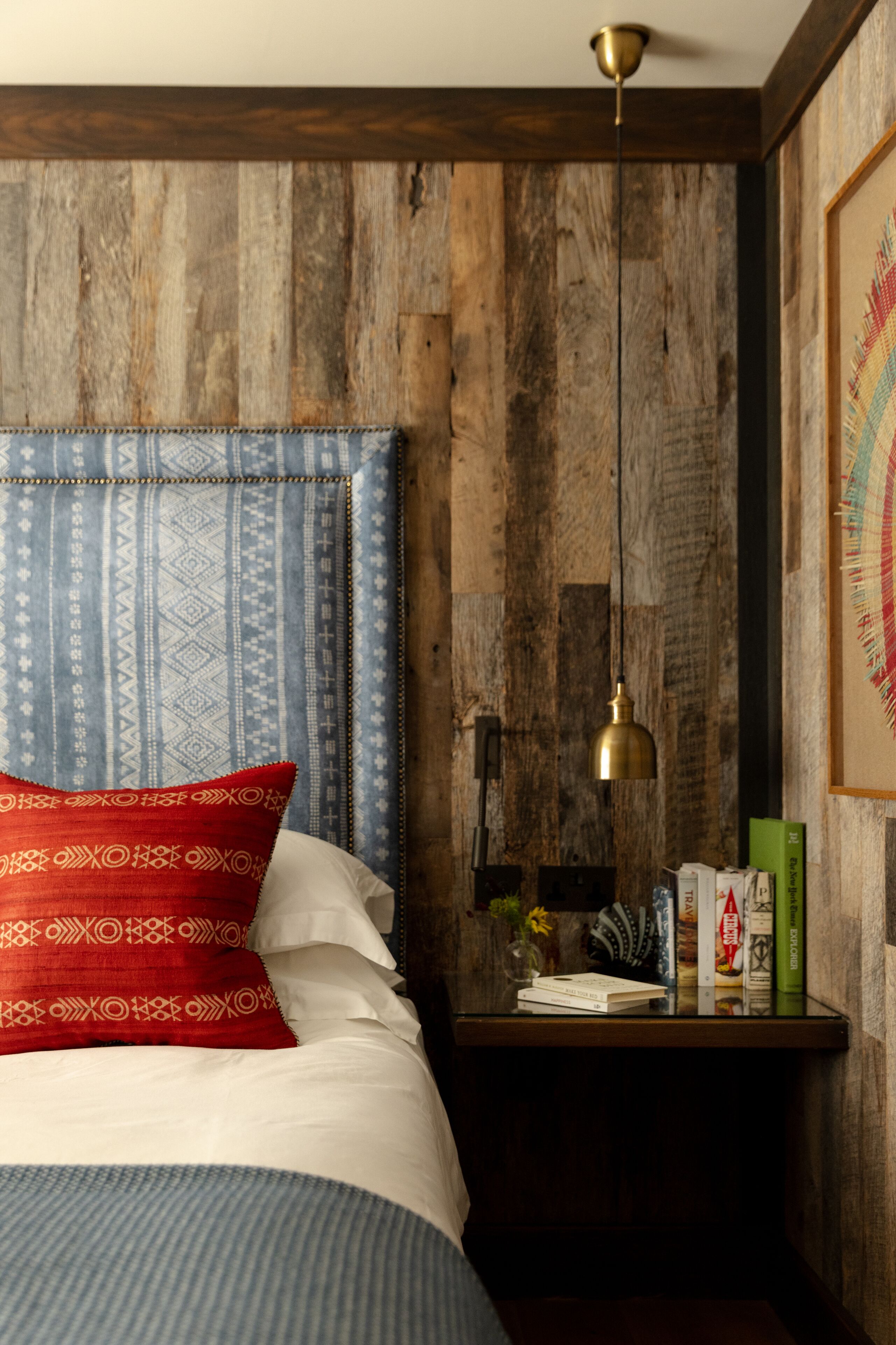 Een slaapkamer met een houten hoofdeinde, een nachtkastje met boeken en een lamp, en een lijst met een afbeelding aan de muur.