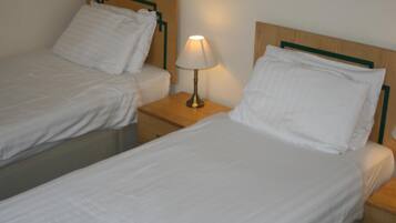 A hotel room with two single beds, each with white linens and pillows, a wooden headboard, and a bedside table with a lamp.