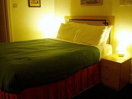 Una cama con colcha verde y almohadas blancas, una mesita de noche con lámpara y cabecera de madera.