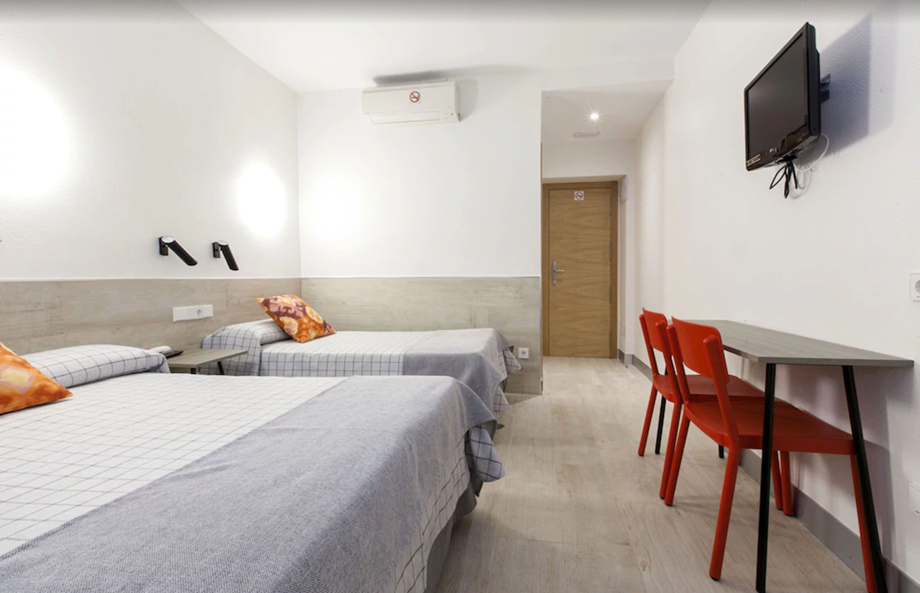 A hotel room with two beds, a desk, a chair, a TV, and an air conditioning unit.