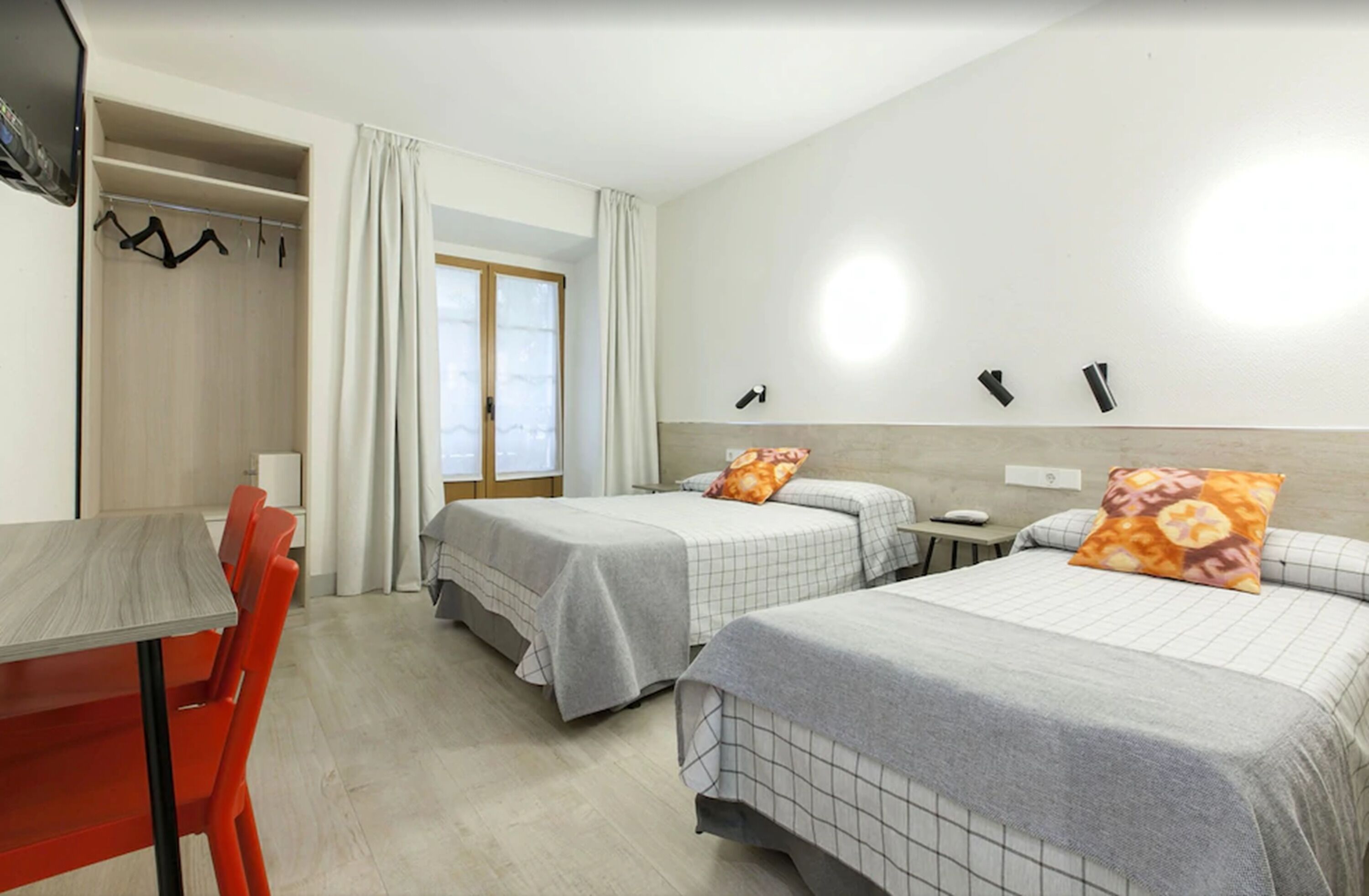 A hotel room with two beds, a table, and chairs. There is a window with curtains, a coat rack, and a wall-mounted light fixture.