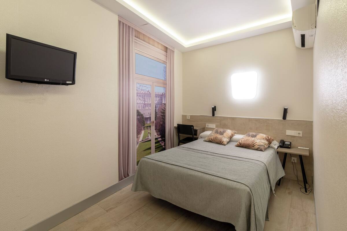 A hotel room with a bed, a flat-screen TV mounted on the wall, a bedside table with a lamp, a phone, and a window with curtains.