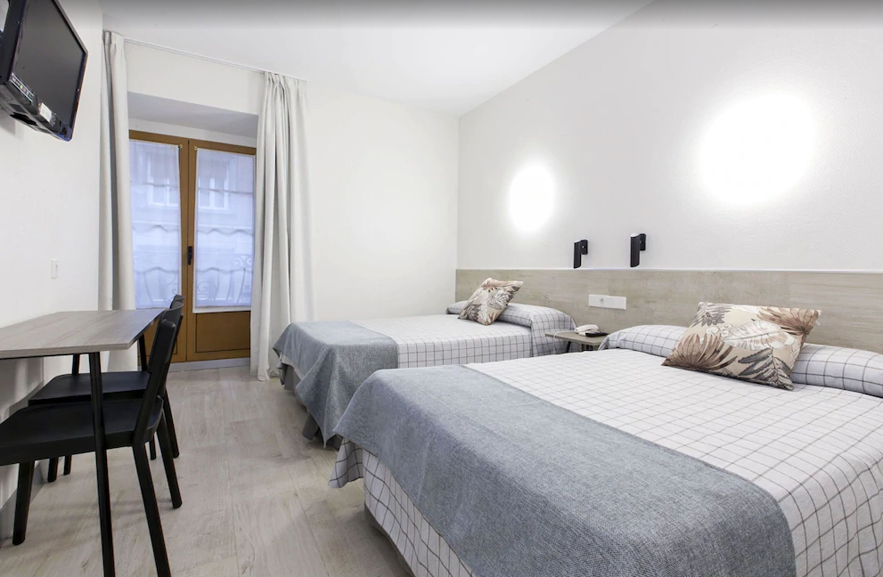 A hotel room with two beds, a desk, and a chair. There is a television mounted on the wall and a window with curtains.