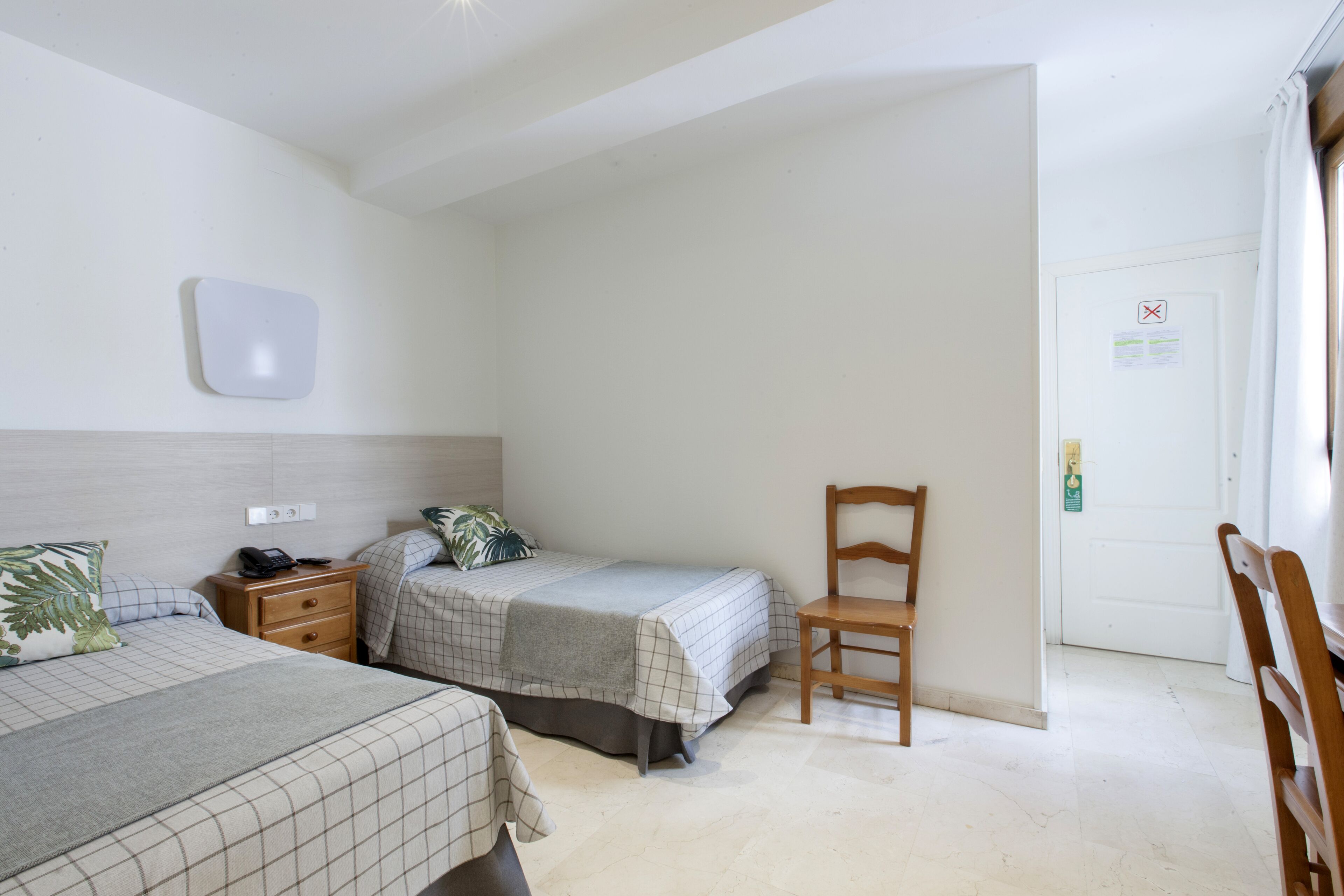 A hotel room with two beds, a wooden chair, a bedside table with a phone, and a wall-mounted light fixture.