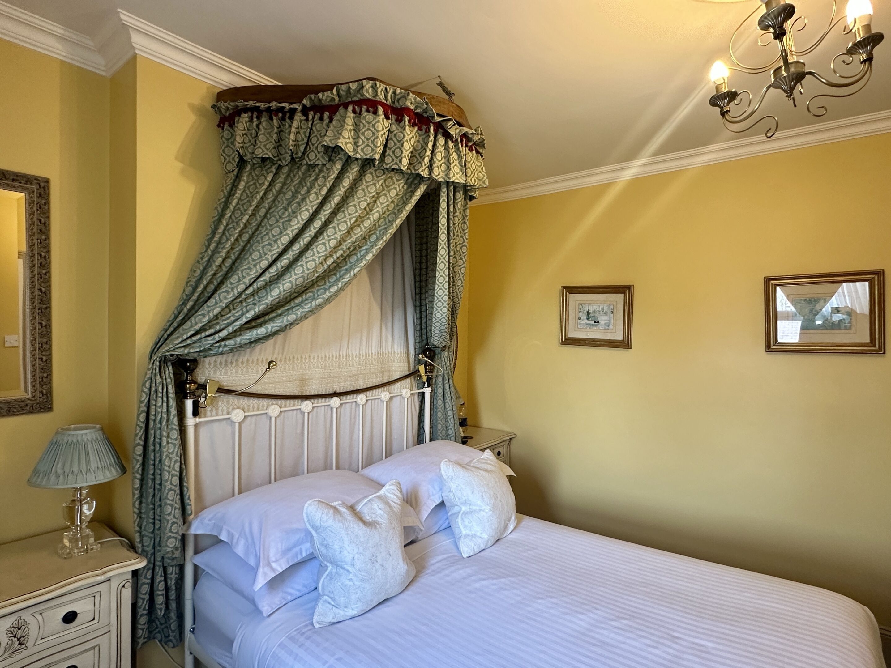 A bedroom with a canopy bed, a bedside table with a lamp, and two framed pictures on the wall.