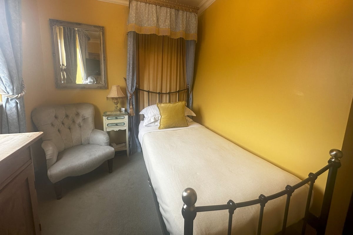 Una habitación de hotel con cama, una silla, una mesita de noche, un espejo y cortinas.