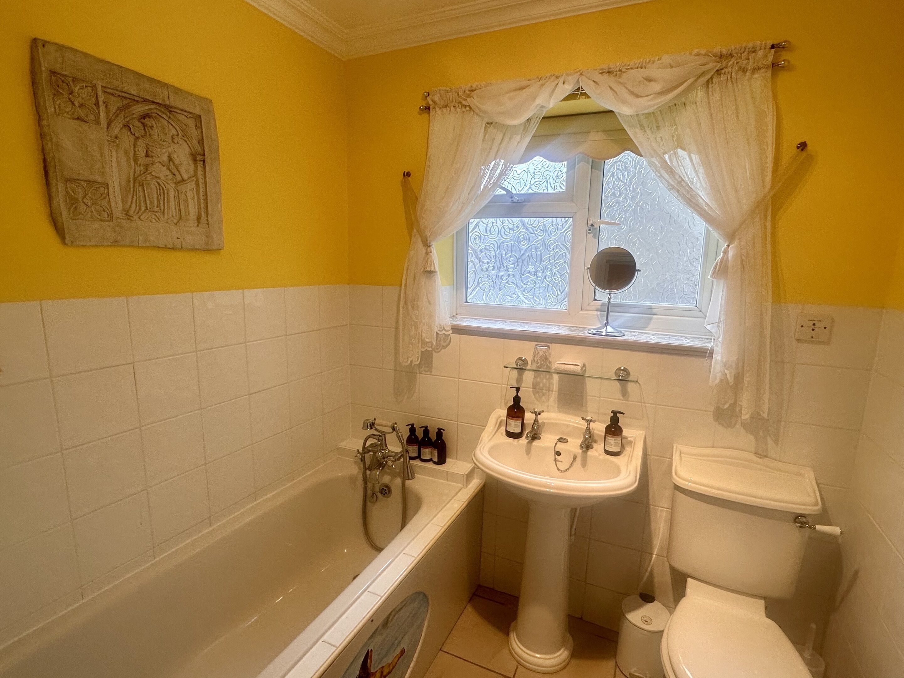 Een badkamer met bad, wastafel en toilet. Er is een raam met gordijn en een spiegel boven de wastafel.