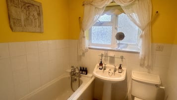Een badkamer met bad, wastafel en toilet. Er is een raam met gordijn en een spiegel boven de wastafel.