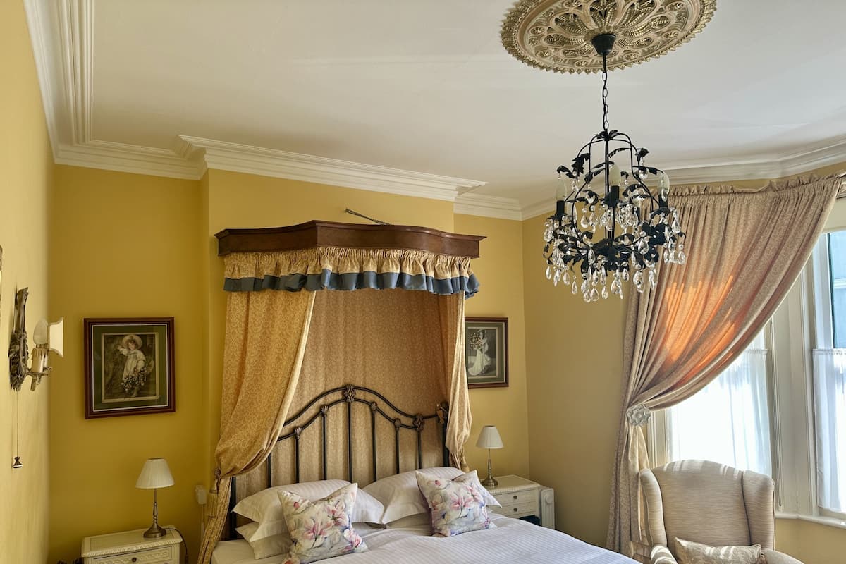 Un dormitorio ordenado con una cama con dosel, una araña y una silla junto a la ventana.