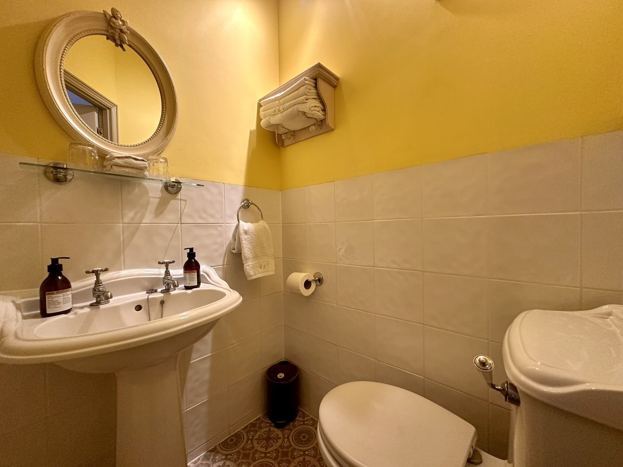 Une salle de bain avec un lavabo blanc, un miroir et des toilettes. On y trouve des articles de toilette sur le lavabo, un porte-serviettes et une poubelle.