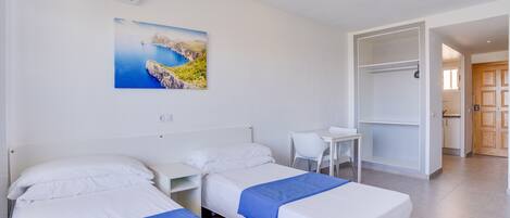 A hotel room with two beds, a desk, a chair, a wardrobe, and a painting of a coastal landscape.