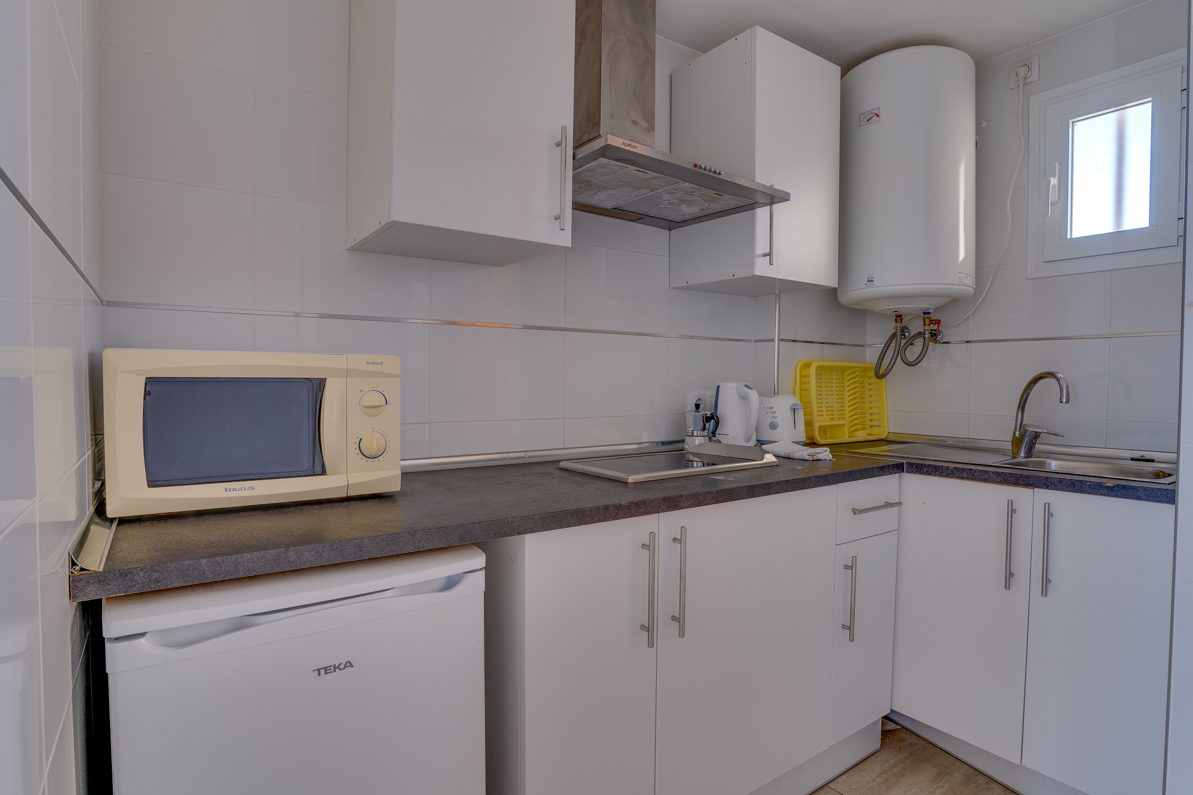 Una cocina compacta con armarios blancos, un microondas integrado, un lavamanos y un calentador de agua.