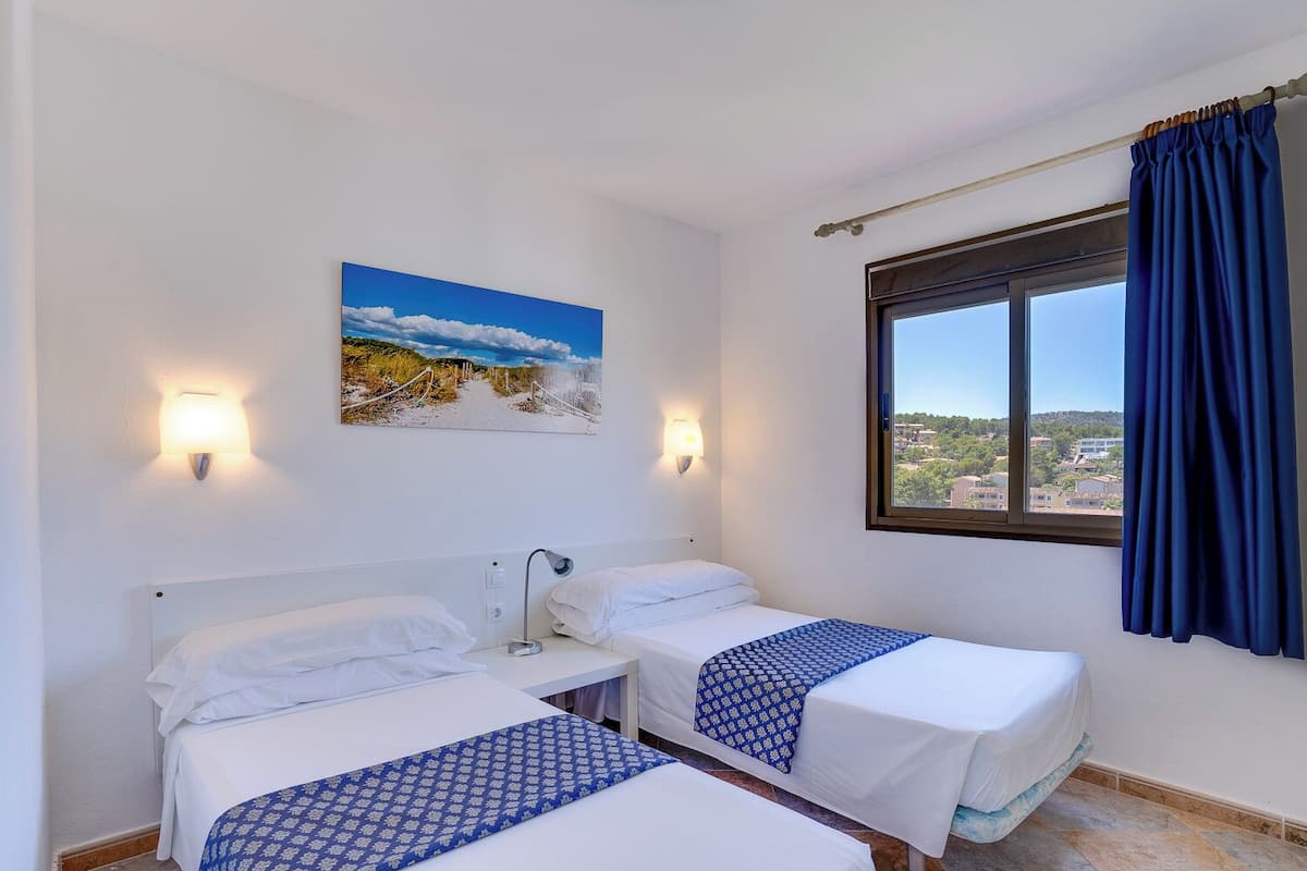 Ein Hotelzimmer mit zwei Betten, einem Strandbild, einem Fenster mit Blick auf Gebäude und einem Vorhang.