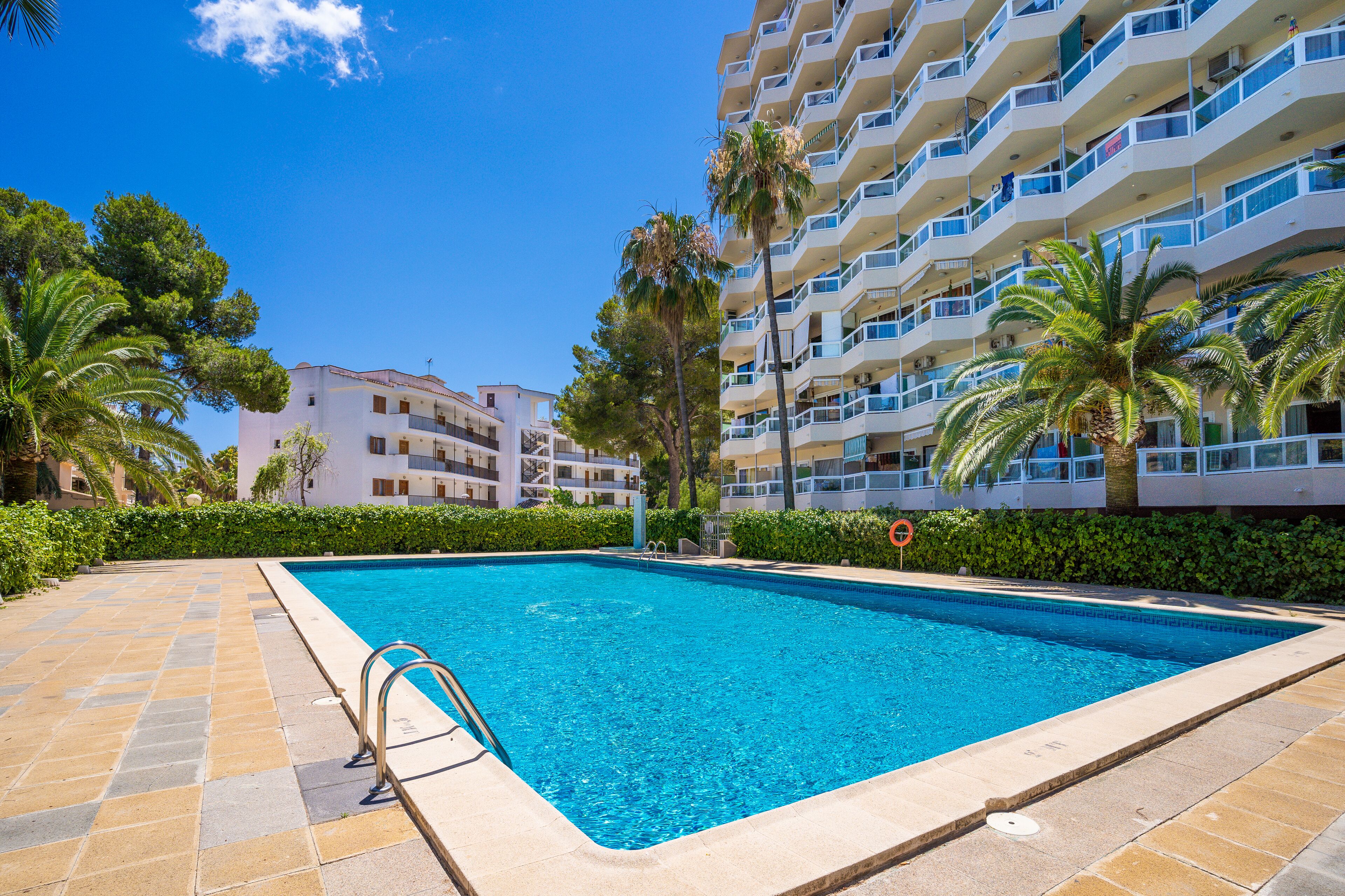 Una piscina con suelo de baldosas, rodeada de palmeras y un edificio de varios pisos con balcones.