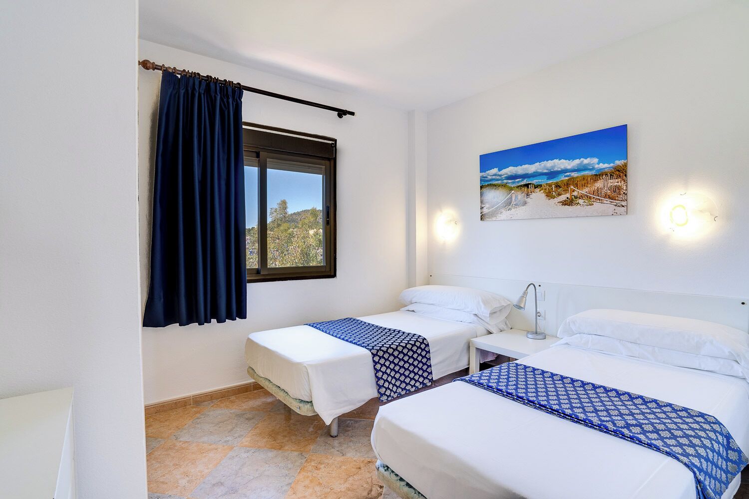 A hotel room with two single beds, a window with a view of trees, a painting on the wall, and a nightstand with a lamp.