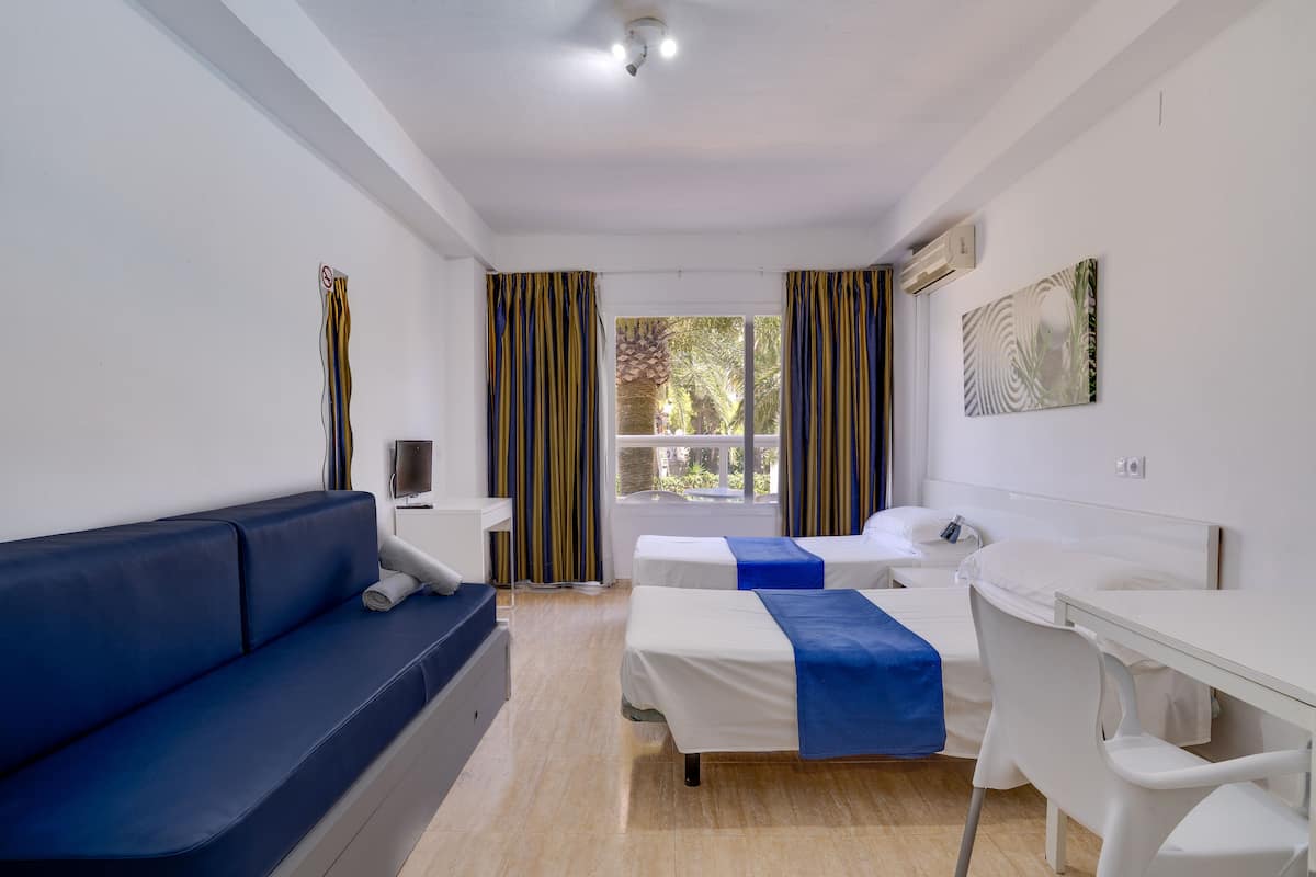 Ein Hotelzimmer mit einem blauen Sofa, zwei Einzelbetten, einem Schreibtisch und einem fenster mit Vorhängen.