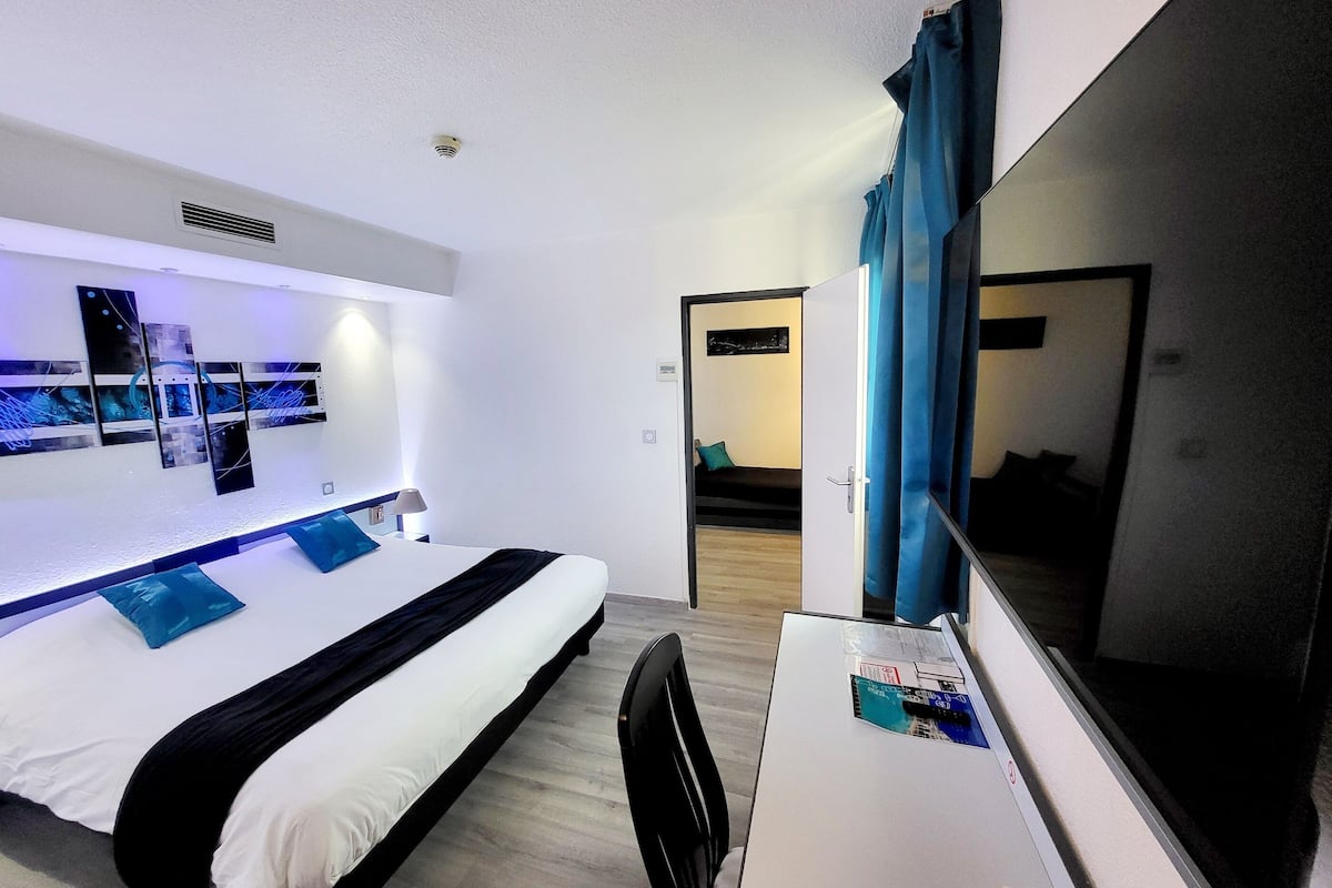 Habitación de hotel con una cama, un escritorio con computadora, un televisor y una puerta que da a otra habitación.