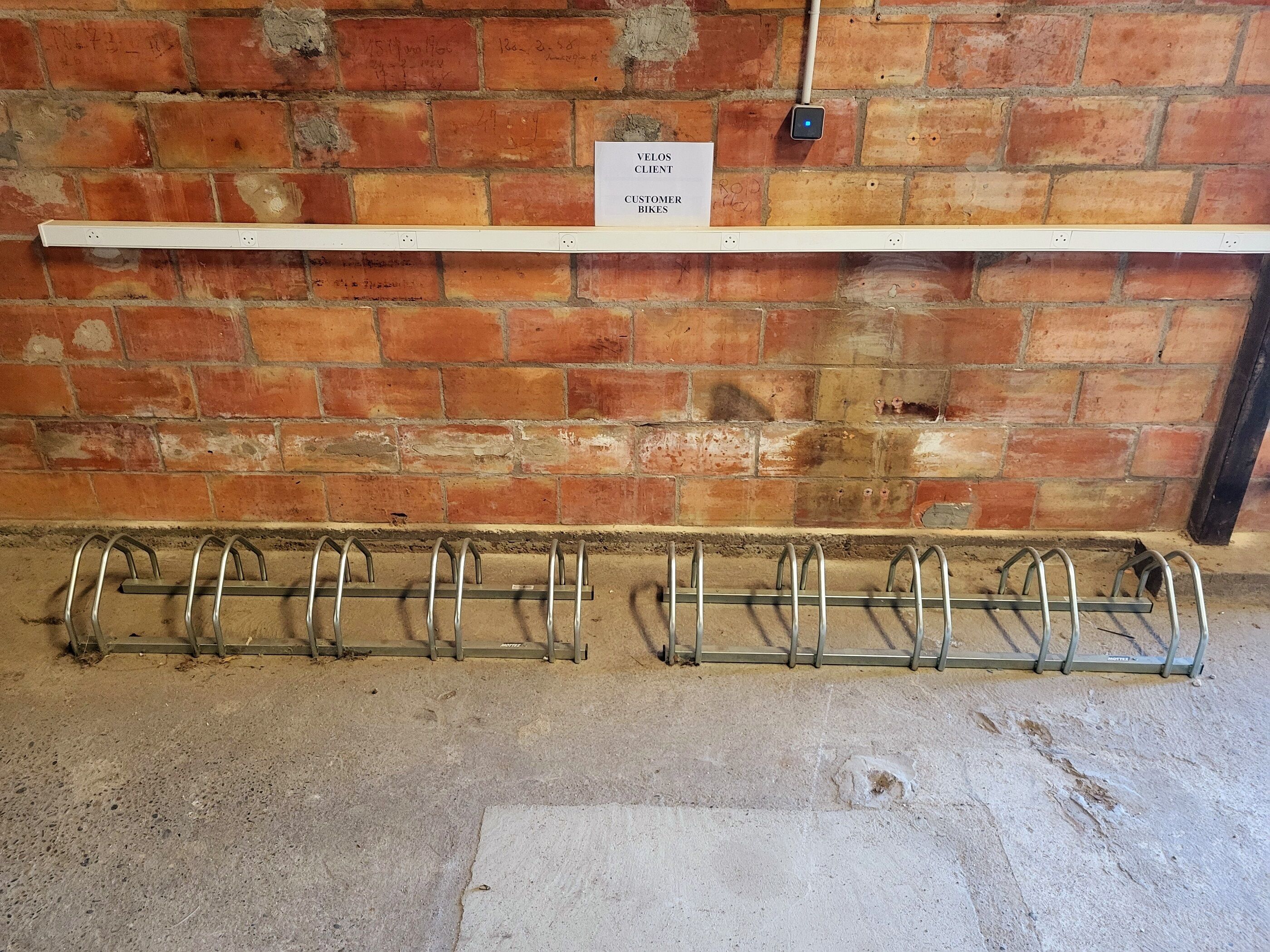 Un muro de ladrillo con un letrero que dice "VELOS CUSTOMER BIKE" y un soporte para bicicletas con varios ganchos.