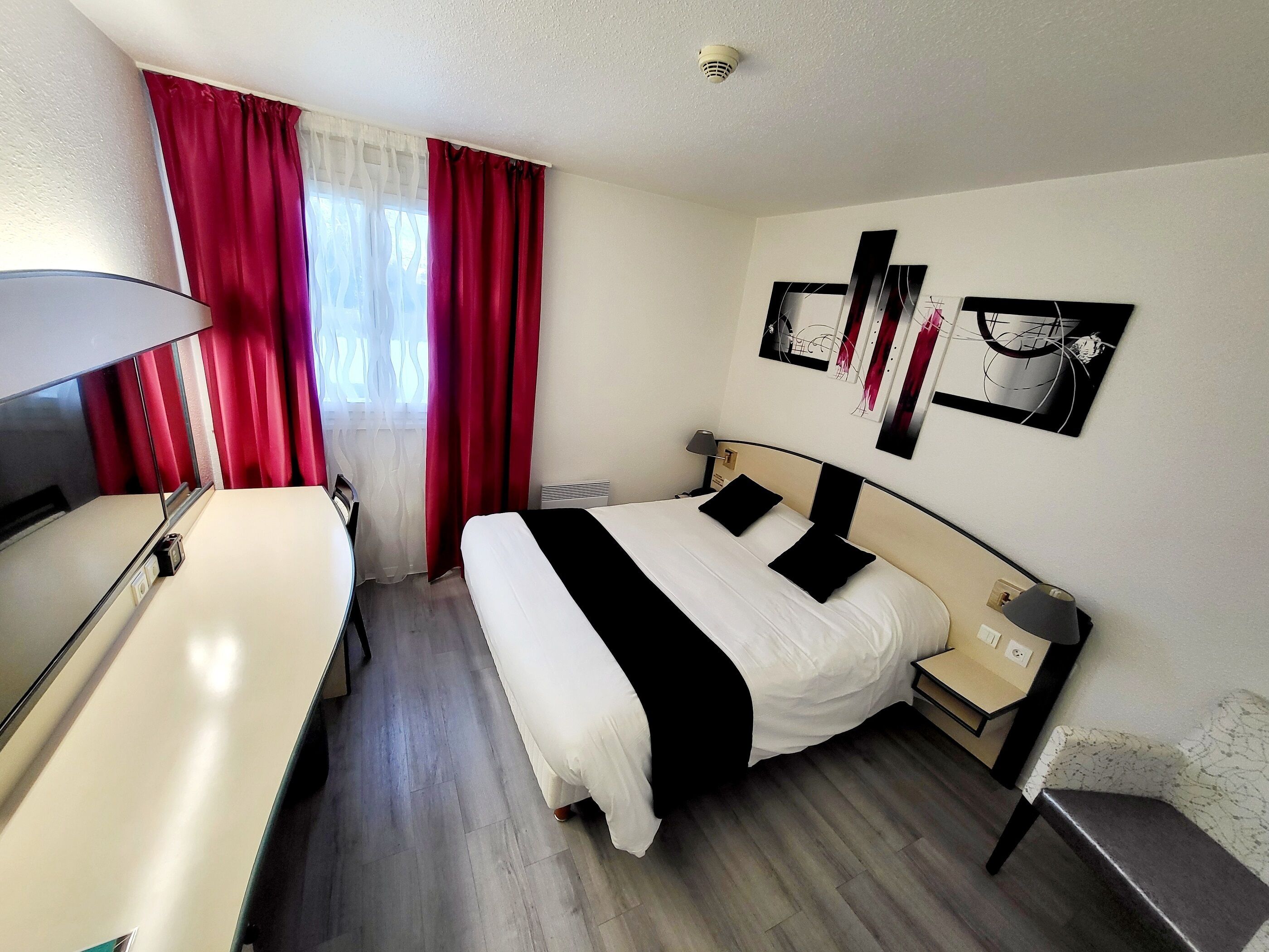 Quarto de hotel com cama, mesa de cabeceira, luminária e escrivaninha com cadeira.