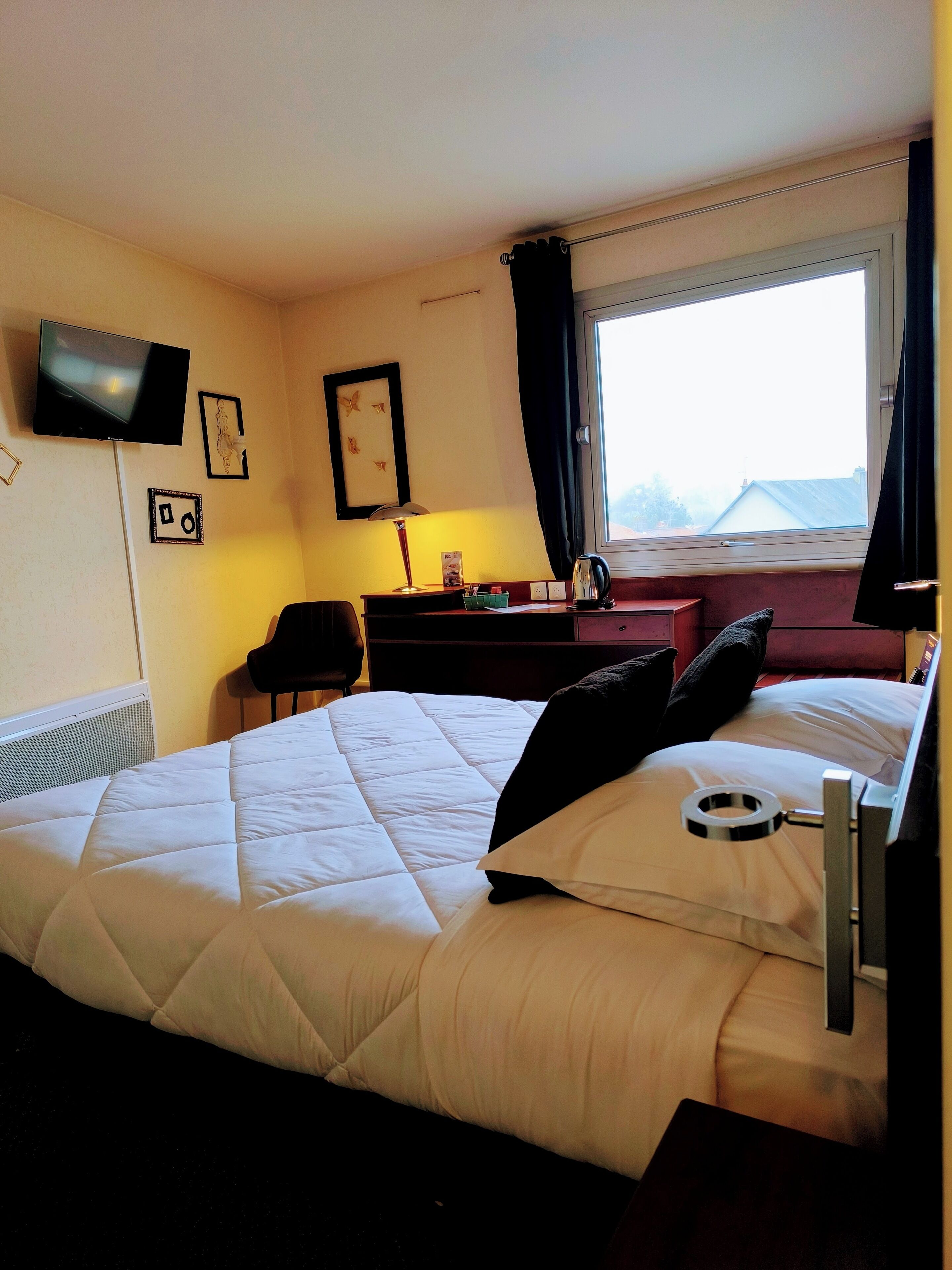 A hotel room with a bed, a desk, a chair, a TV mounted on the wall, and a window with curtains.